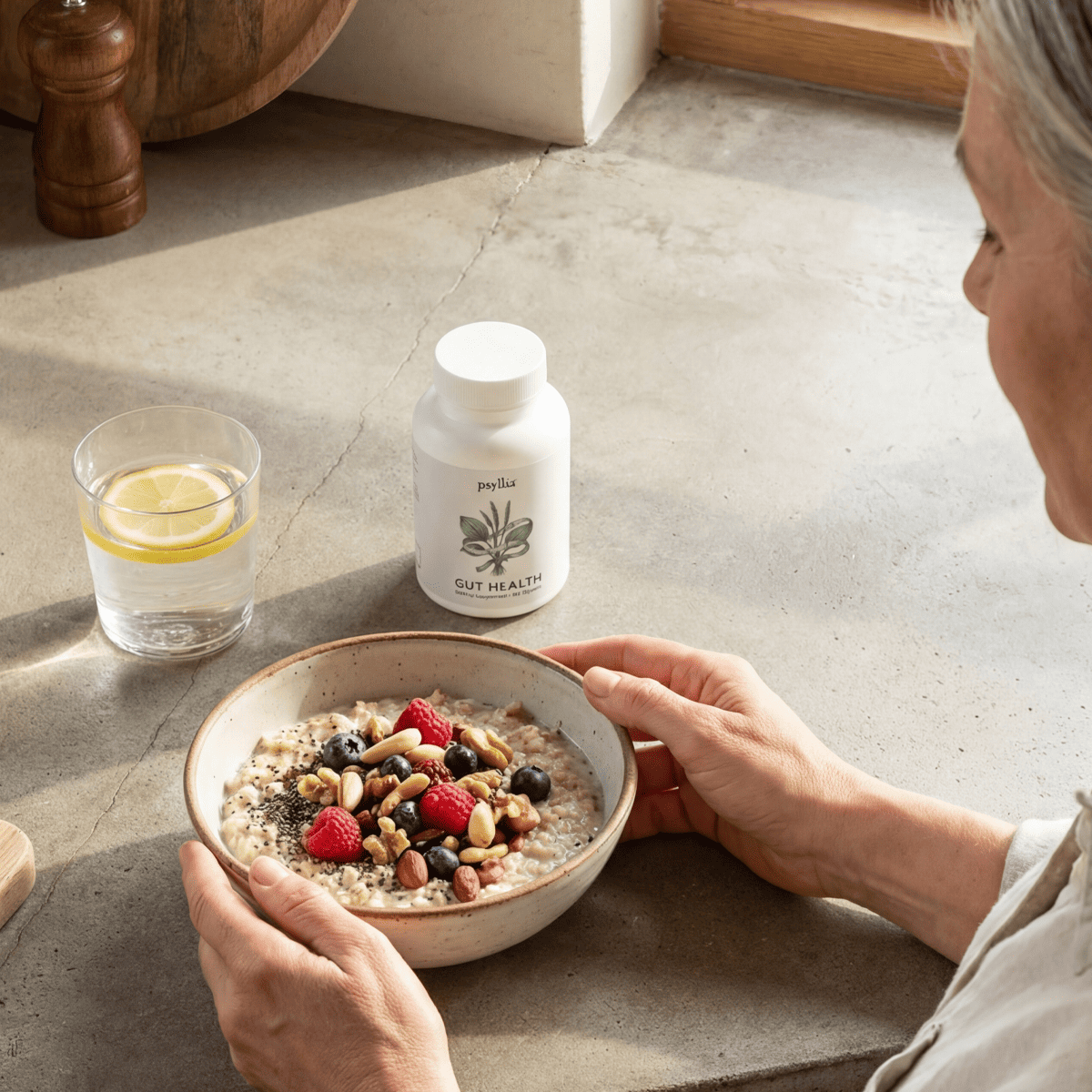 A bright kitchen counter scene: bottle of Psyllia next to a glass of lemon water and a bowl of oats with berries and nuts
